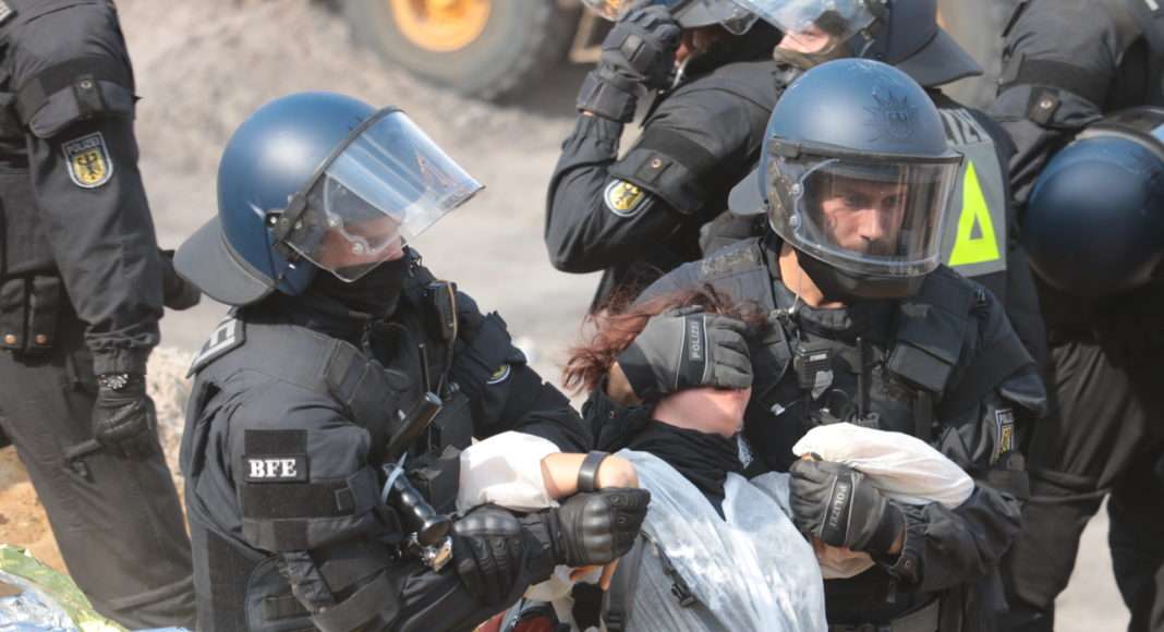 Après plusieurs heures en plein soleil, les policiers entament les arrestations. Celles et ceux qui ne suivent pas docilement se voient poussés, fortement maintenus au visage ou les poignets tordus pour les faire avancer.